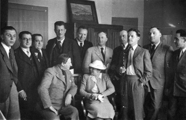 Motiv: Kirkeby siddende i samtale med en dame omgivet af 10 stående herrer